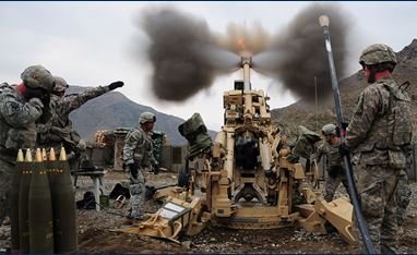 A group of soldiers stand around a tan machine, which is in the process of firing an explosive. There are mountains in the background, and ammunitions set up beside the machine. 