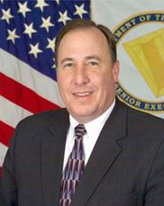 Image of a man (John Hedderich, III) seated in front of the American Flag. He wears a gray suit and has a muti-colored, red and blue tie on. He has blue eyes. 
