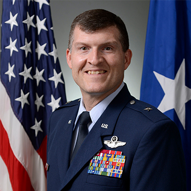 Image of Brig Gen Hinote, USAF. Brig Gen is in a military uniform and stands before the American flag, smiling at the viewer. He has short cropped hair. 