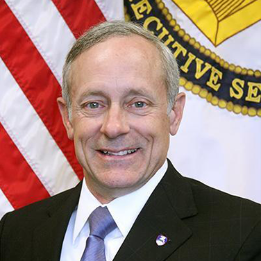 Image of Richard De Fatta, a Caucasian male. He is seated before a U.S. flag and is gazing at the camera with a smile on his face. He wears a dark suit and has a purple tie. 