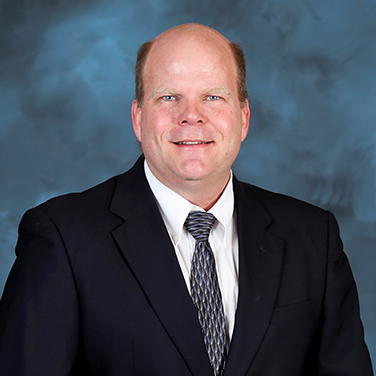 Image of Dr. Lonnie Love, a middle aged Caucasian man with blue eyes and a grin on his face. He is seated before a blue backdrop and wears a dark jacket and blue-gray tie. 
