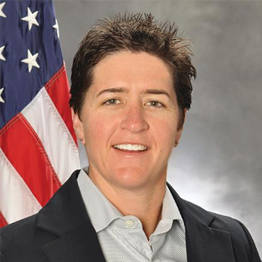 Image of Adele Ratcliff. A woman with short brown hair stands in front of an American flag and a gray background. She has a dark blazer on and a gray shirt. She has an open mouth smile. 