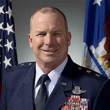 Image of Maj Gen Mike Fantini. Fantini is in front of the American and USAF flags in his blue military uniform. She has an open mouth smile and numerous badges and insignia on his uniform. 