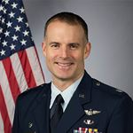 Image of Lt Col Brian Stiles, USAF. Stiles is seated before an American flag. He has an open mouth smile, has short hair, and is wearing an Air Force uniform with buttons and insignia. 