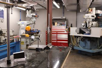 Image of SOFWERX's Foundry; a large open room with concrete floors and large machinery. In front of the viewer is a table which sits a scale and other test instruments. 