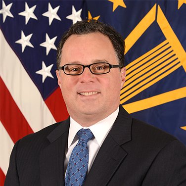 Image of Robert Gold; Mr. Gold is seated before a American flag. He wears a dark suit, blue tie, and has black-rimmed glasses and is smiling with his teeth showing. 