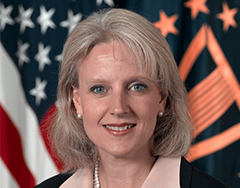 Image of woman with light hair, blue eyes, and an open mouth smile. She is wearing pearl earrings and necklace. She sits before the U.S. Flag. 
