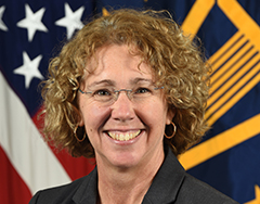 Image of Sandy Magnus, who is seated before the U.S. flag. Magnus has curly blond-brown hair, glasses, hop gold earrings, and is smiling with her mouth open. 