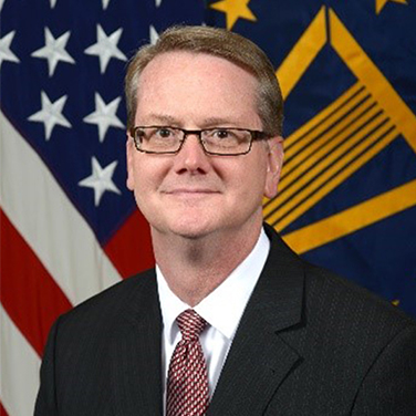 Image of David Cadman. Cadman sits in front of a U.S. flag. He wears a black suit with a maroon tie. He has light hair, wears classes, and has a closed-mouth smile. 