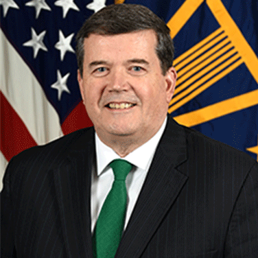 Image of Dr. Raymond O’Toole; O'Toole is seated in front of an American and military flag. He has a dark suit and green tie. He has dark hair and is smiling showing his teeth. 