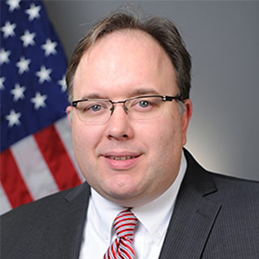 Image of Neil Graf; Graf sits in front of a U.S. flag. he is wearing a gray suit and has a red and white striped tie. He wear glasses, has light brown hair and is giving a small smile. 