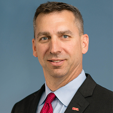 Image of Wes Hallman; Hallman is standing before a blue background, he has a dark suit and red tie. He has light-brown hair and wears an NDIA pin. 