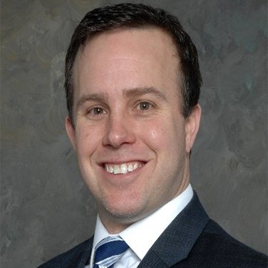 Image of Kevin McGrath. McGrath is seated before a gray background. He has a dark suit and blue striped tie. He has dark hair and has an open mouth smile. 