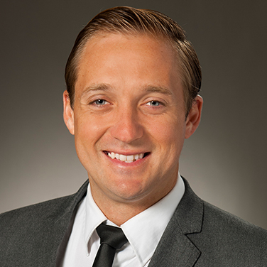 Image of David Rampton; Rampton sits before a gray background. He wears a gray suit and tie, is smiling with his teeth showing, and light hair parted on the side. 