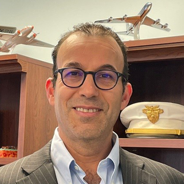Image of Moshe Schwartz. Schwartz stands in front of a bookshelf with a military hat and model airplanes. He wears a light suit with a blue shirt and dark, rounded glasses. He smiles with his teeth showing. 