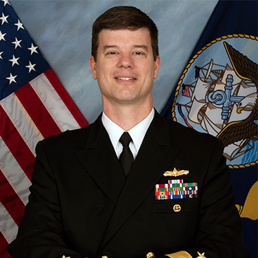 Image of RDML Kevin Byrne, USN who is seated in front of the U.S. and USN flags. He is wearing his navy uniform. 