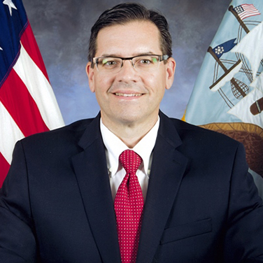 Image of Don Hoffer, seated in front of the U.S. and U.S. Navy flags; he wears a dark navy suit and a red tie. He has glasses and is smiling with his teeth showing. 