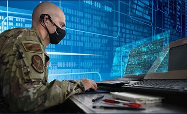 Image of a man in military attire sitting at a computer; he is wearing a face mask.